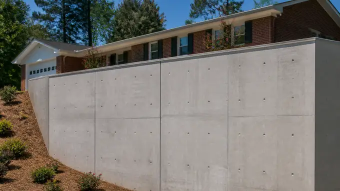 Tall poured concrete basement wall with snap-tie holes visible at a red-brick ranch home in Sandy Springs, GA, in clear late-morning sun.